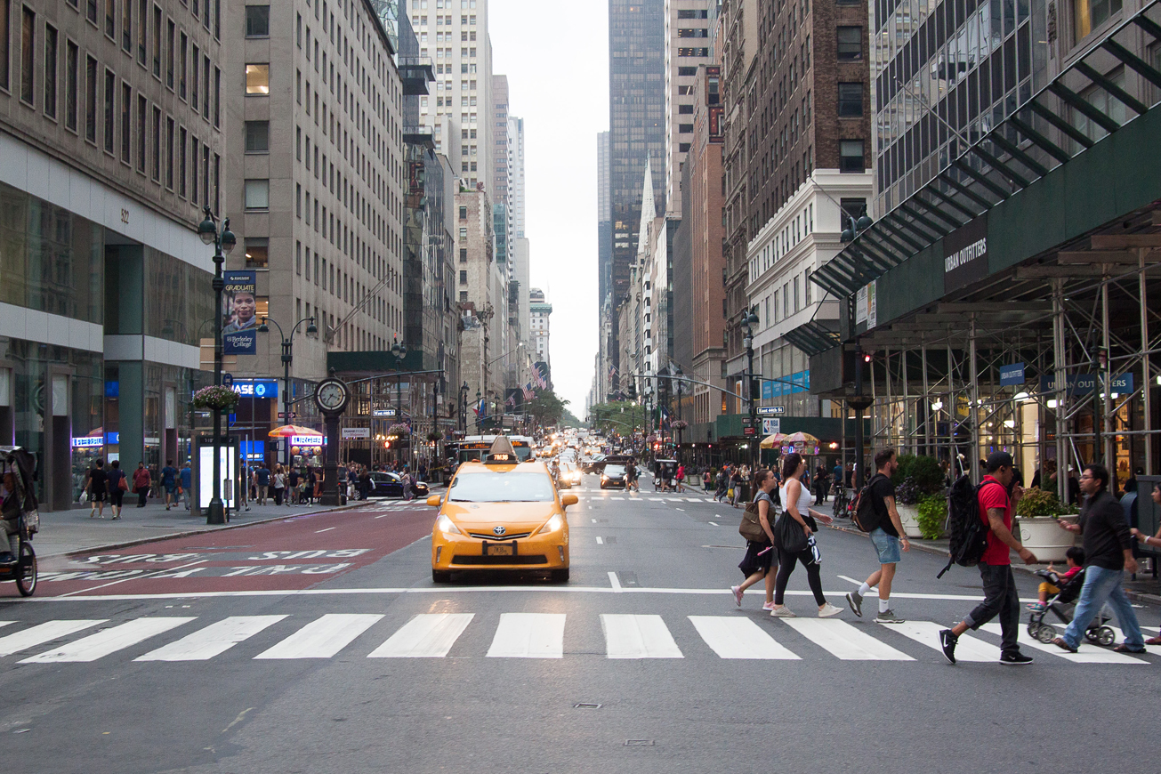 An intersection in NYC with a yellow taxi stopped at a light, and five pedestrians crossing the street. 